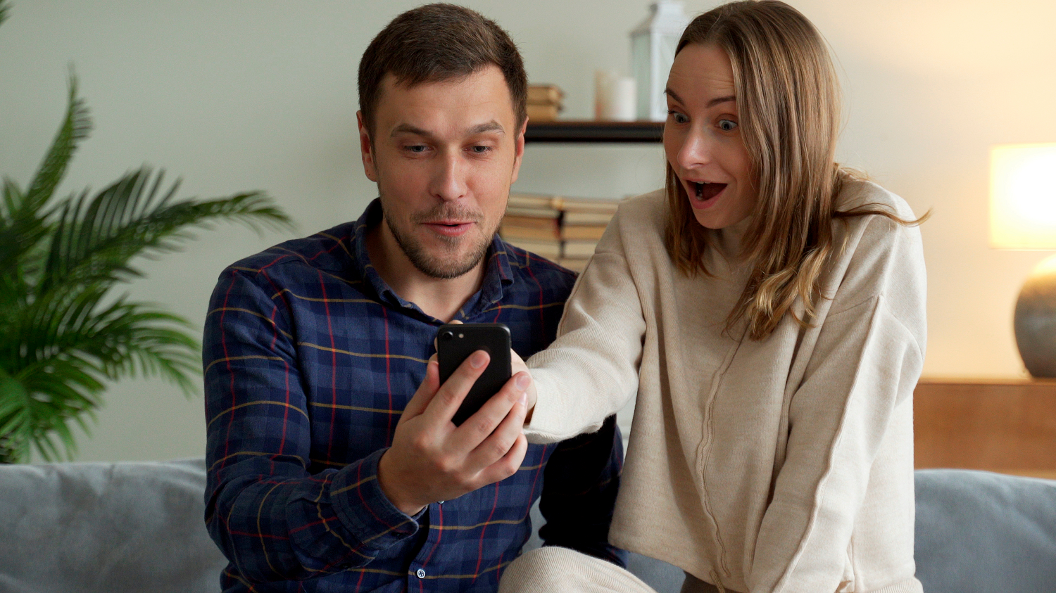A young couple at home, gasping as they have won the lottery on their phone, representing the impact of using a charity lottery service