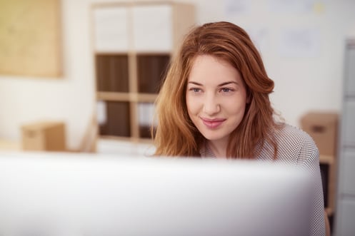 Attractive young woman working on a desktop computer smiling as she leans forwards reading data analysis results to aid her decision making, view over the monitor