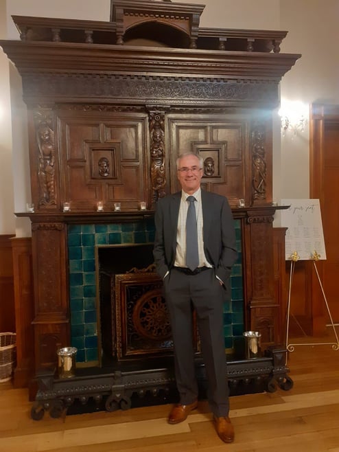 Steve Hubbard standing in front of a grand fireplace. Steve is wearing a grey suit and grey tie.