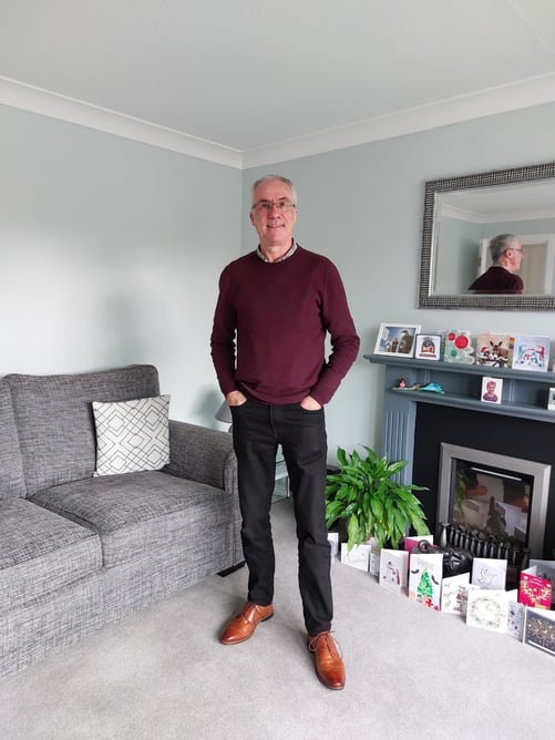 Steve Hubbard standing in a living room, wearing a red jumper, black trousers and brown shoes. In the room is a fireplace with Christmas cards, a sofa and mirror.