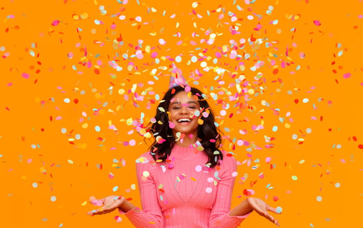 young woman in pink long sleeved top with arms outstrethced catching confetti against an orange background