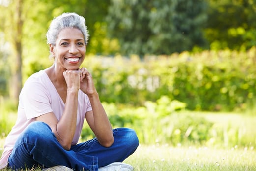 older black lady with grey hair in t-shirt and jeans sitting cross legged on the grass and smiling on a summers day