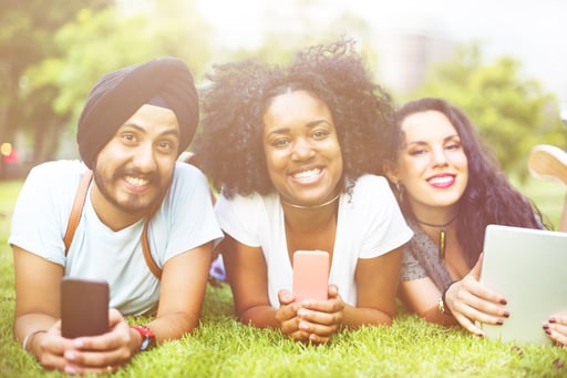 three multi racial young people laying on the grass smiling at the camera, with smart phones or tablet in hands